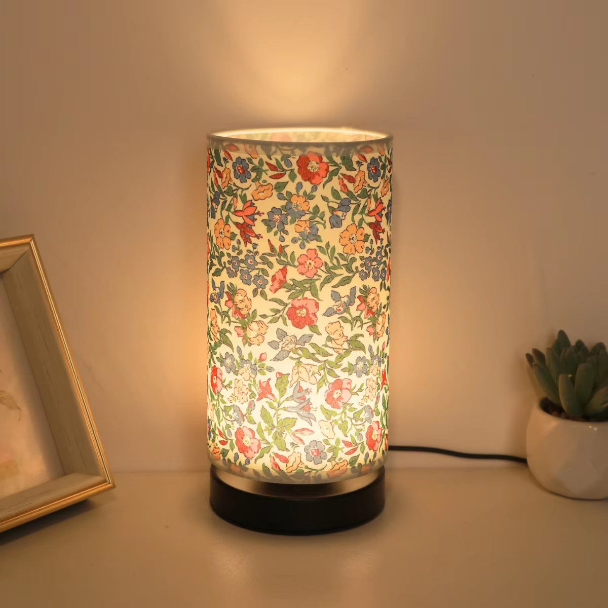 Floral fabric bedside lamp glowing warmly on nightstand with framed photo and small plant, cozy decorative lighting for bedroom ambiance