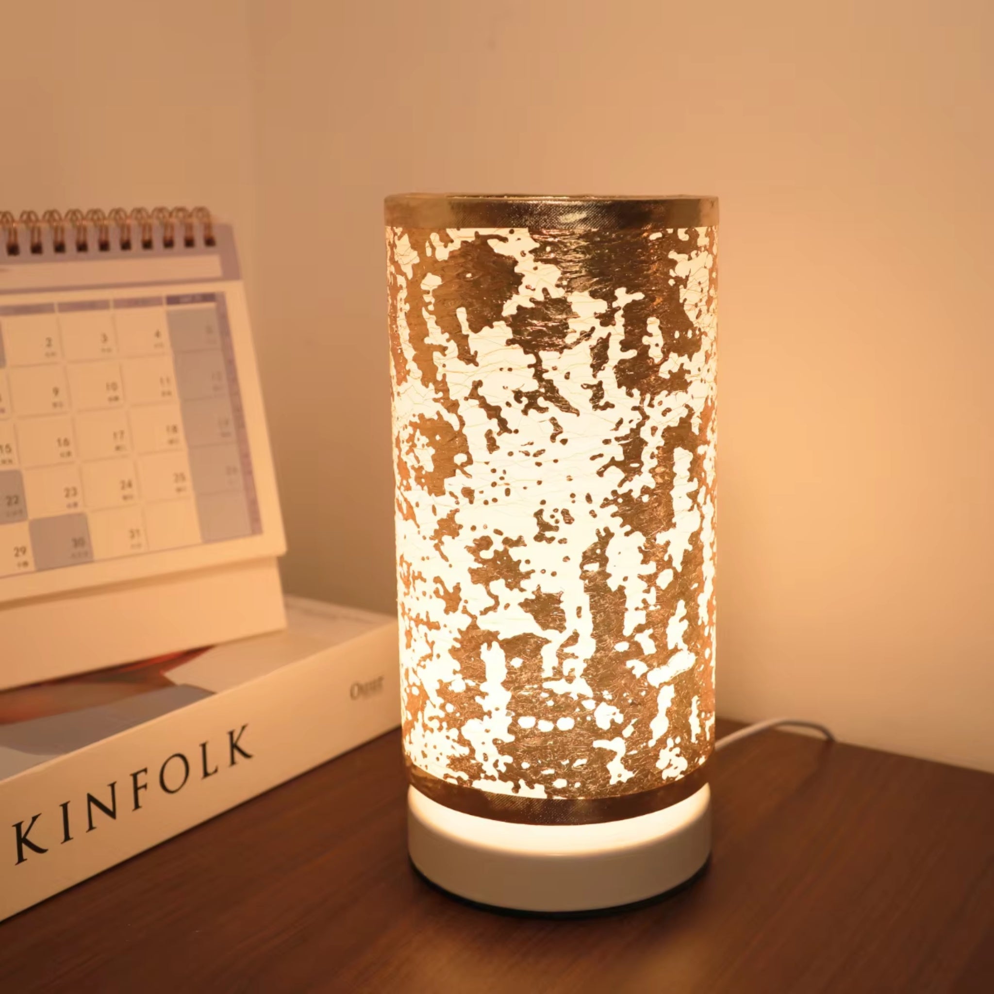 Gold textured bedside lamp glowing warmly on wooden table with calendar and books, modern decorative ambient lighting for bedroom or desk