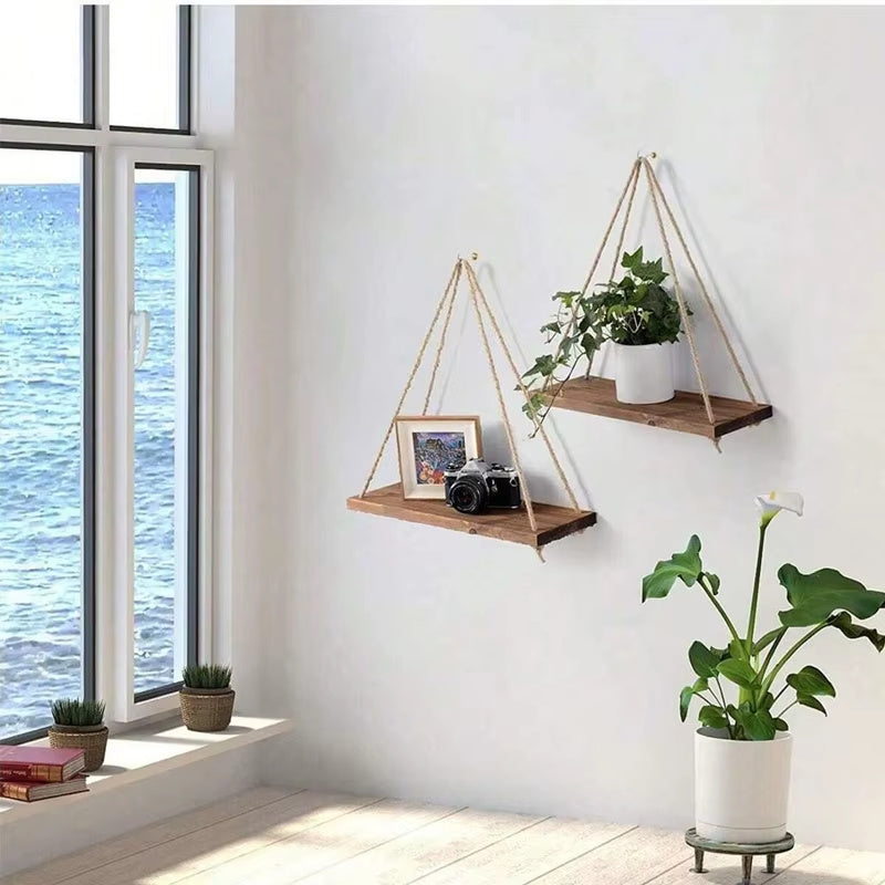 Modern wood rope hanging wall shelves set displayed in a bright room holding a potted plant camera and framed photo mounted on a white wall near a window