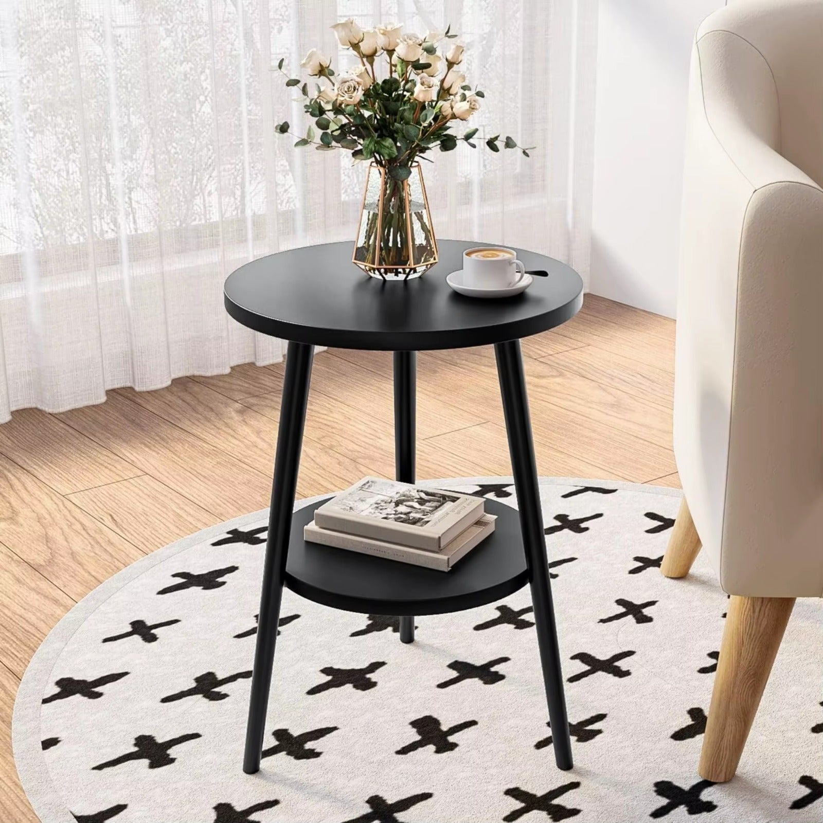Black round side table with a vase of flowers and a cup on a white rug with black patterns, next to a beige armchair.